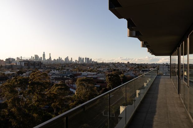 A wide elevated balcony or terrace with a glass balustrade and dark projecting roof canopy, overlooking a dense urban neighbourhood and distant high-rise city skyline. Full-height glazing lines the building facade on the right, while warm late-afternoon light creates long shadows and expansive metropolitan views.