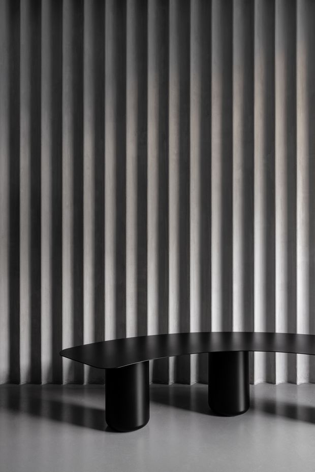 A monochromatic, minimalist interior scene featuring a sculptural black curved table with two cylindrical pedestal legs, set against a dramatic floor-to-ceiling vertically ribbed or pleated wall treatment. The polished grey floor reflects the table and strong vertical shadows, creating a refined, high-contrast product-oriented composition.
