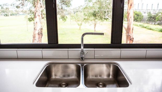 Close-up view of a contemporary kitchen sink area beneath a large black-framed window. The image features a double-bowl stainless-steel undermount sink, chrome gooseneck mixer tap, white tiled splashback with dark grout, and a light glossy benchtop. A leafy rural landscape is visible through the glazing.