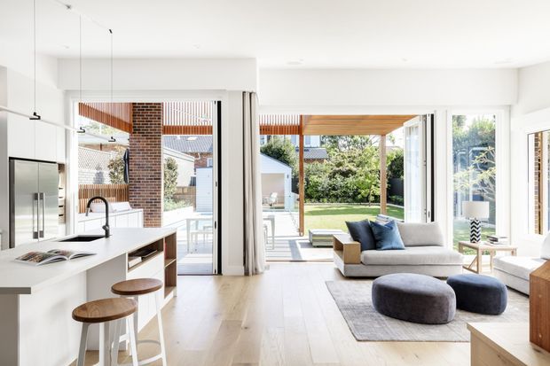 Bright open-plan contemporary living and kitchen area with white cabinetry, a large island and timber stools opening directly onto a landscaped backyard through wide glazed doors. The living zone contains a pale sofa, blue cushions, dark rounded ottomans and a large neutral rug. Exposed brick and timber details add warmth, while abundant glazing creates an airy indoor-outdoor connection.
