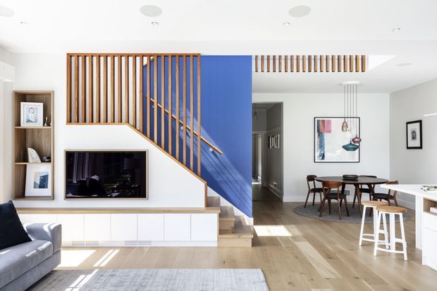 Bright open-plan interior showing a living room, dining area, and kitchen zone connected by a central staircase. The staircase features warm timber vertical balusters and a vivid blue painted wall, with integrated white living-room cabinetry and television below. Pale timber flooring, white walls, contemporary pendant lighting, and a dining table with dark timber chairs create a clean, colourful contemporary family home.