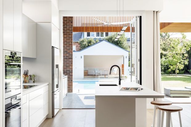 Bright, contemporary open-plan kitchen viewed toward a glazed opening and outdoor pool courtyard. The kitchen features tall white cabinetry, integrated appliances, a minimalist island with a dark inset cooktop or sink, and two timber-topped stools. White-framed glazing and a covered outdoor area connect the kitchen to a landscaped backyard and pool. Warm timber battens and exposed brick add texture beyond the kitchen.