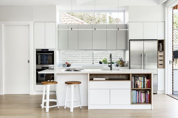 Bright contemporary kitchen with a large white island, integrated cabinetry, stainless-steel refrigerator and ovens, white subway-tile splashback, timber-look flooring, and two timber-topped stools. Full-height glazing and a high-level window bring in abundant natural light, while open shelving displays cookbooks and accessories.