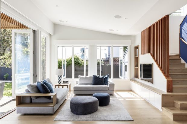 Bright contemporary living room with large glazed doors opening to a garden, pale timber flooring, neutral upholstered sofas, dark blue cushions and ottomans, and a built-in fireplace beneath a sculptural timber slatted stair balustrade. The room has abundant natural light, recessed ceiling lighting, open shelving, and a light, airy layout.