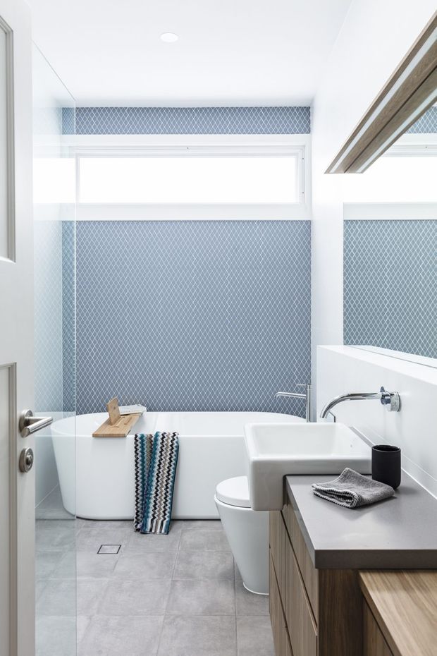 Bright contemporary bathroom with a freestanding white bathtub, wall-mounted chrome fixtures, a white vessel basin, timber vanity cabinetry, large grey floor tiles, and a blue-grey geometric tiled feature wall. A frameless glass shower screen and large mirror contribute to the clean, light-filled interior.