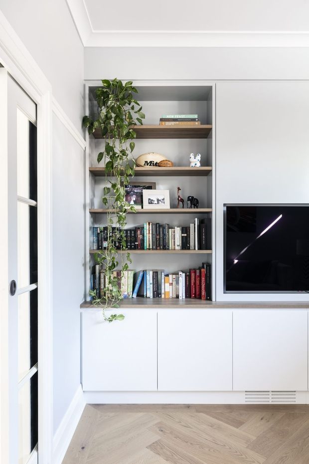 Bright, refined built-in media and library wall featuring open timber shelves filled with books and small decorative objects, white closed cabinetry below, and a wall-mounted black television to the right. A trailing indoor plant adds greenery. The room has white walls and detailed ceiling cornices, a timber-look herringbone floor, and a glazed white internal door at left.