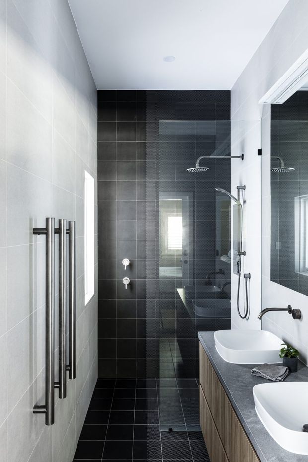 A narrow, contemporary bathroom with a walk-in glass shower at the rear, dark charcoal tiled feature walls and floor, pale wall tiles, a wall-mounted rainfall shower and handheld shower, and a long timber-look vanity with two white vessel basins and a grey benchtop. A large mirror reflects the vanity and shower, while a vertical heated towel rail is mounted on the left wall.
