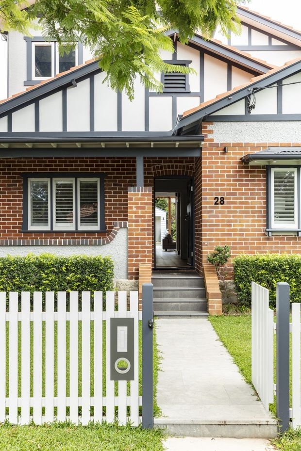Front exterior view of a compact multi-level residential townhouse. The facade combines red brickwork with white rendered upper walls framed by dark timber-like battens, steeply pitched intersecting roofs, projecting eaves, and louvred ventilation openings. A white picket fence, manicured hedges, paved path, and recessed entry porch lead to the front door. The house number 28 is visible beside the entrance.