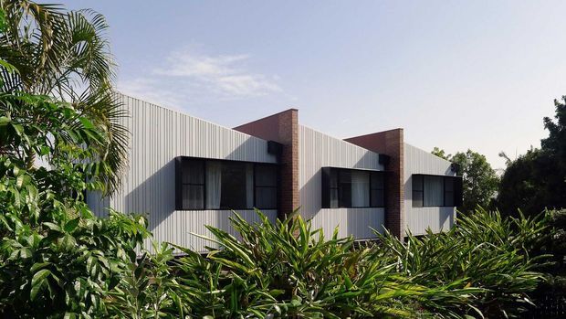 Contemporary low-rise building facade with repeated projecting brick elements, vertically ribbed light-coloured cladding, and dark-framed windows, partly screened by dense tropical landscaping.