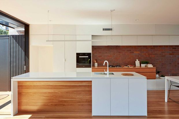 Bright contemporary kitchen with a large white island featuring a warm timber-panelled base and integrated sink. White full-height cabinetry, timber flooring, and a red exposed-brick splashback define the space, with black-framed glazing and a glimpse of a dining area at right.