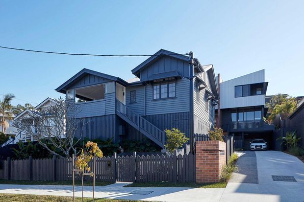 Street-facing view of a multi-level residential property with dark blue-grey horizontal cladding, pitched gabled roof forms, external stairs, a timber picket fence, brick entry pillar, and a concrete driveway leading to a garage. A contrasting contemporary flat-roof extension is visible to the right.