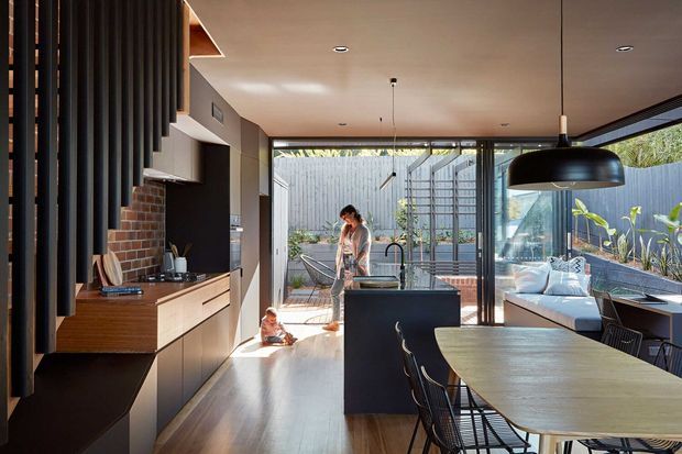 Contemporary open-plan kitchen and dining area with a dark island, timber cabinetry, brick splashback, black pendant lighting, and a long dining table. Full-height glazing opens toward a landscaped outdoor area and brings in abundant natural light.