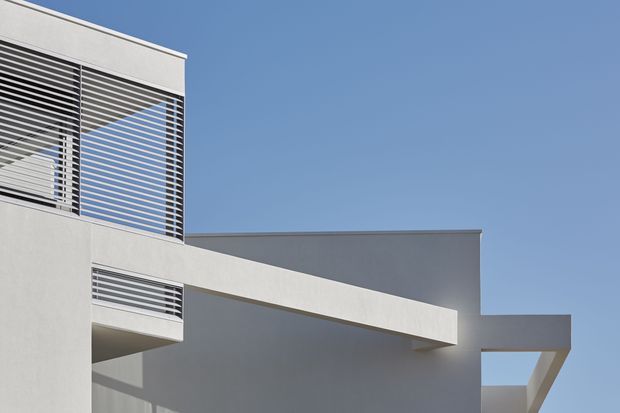 A close-up exterior view of a minimalist modern building under a clear blue sky. Smooth pale rendered walls form strong rectilinear volumes, with projecting slab-like forms and recessed areas. Horizontal metal louvres screen the upper-level glazing and balcony openings, creating crisp linear shadows and a clean geometric facade.