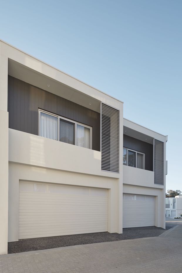 Exterior photograph of a contemporary multi-storey attached residential building, likely townhouses, with clean white rendered forms, dark vertical cladding, recessed upper-level balconies, large louvred privacy screens, glazed doors and windows, and integrated white sectional garage doors. The building fronts a paved driveway or shared access lane under a clear sky.