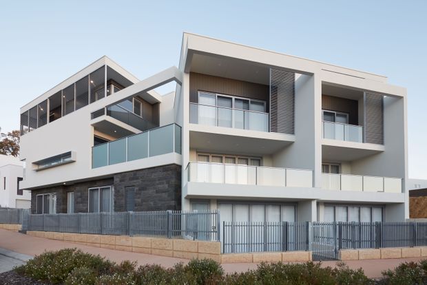 Exterior view of a contemporary multi-storey residential apartment or townhouse development. The building features crisp white rendered volumes, projecting rectangular balconies with clear glass balustrades, dark stacked-stone cladding at lower level, vertical metal privacy screens, and a dark metal perimeter fence. The facade has a clean geometric composition with flat roof forms and large glazed openings.