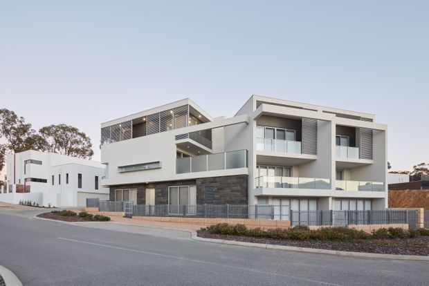 A street-level exterior photograph of a contemporary multi-storey residential building, likely a townhouse or apartment development. The facade combines white rendered volumes, dark stone cladding, extensive glazing, recessed balconies with glass balustrades, horizontal louvres, and a flat roofline. Adjacent modern homes and a paved street are visible in the suburban setting.
