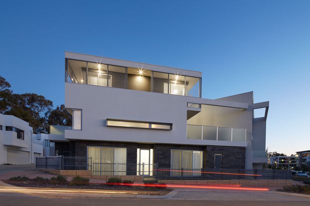 Contemporary multi-storey residential facade photographed at dusk, featuring a box-like flat-roof form, pale rendered upper walls, dark masonry cladding at ground level, extensive upper-level glazing with horizontal louvres, and a projecting covered balcony or terrace. The building fronts a paved street and is softly illuminated from within.