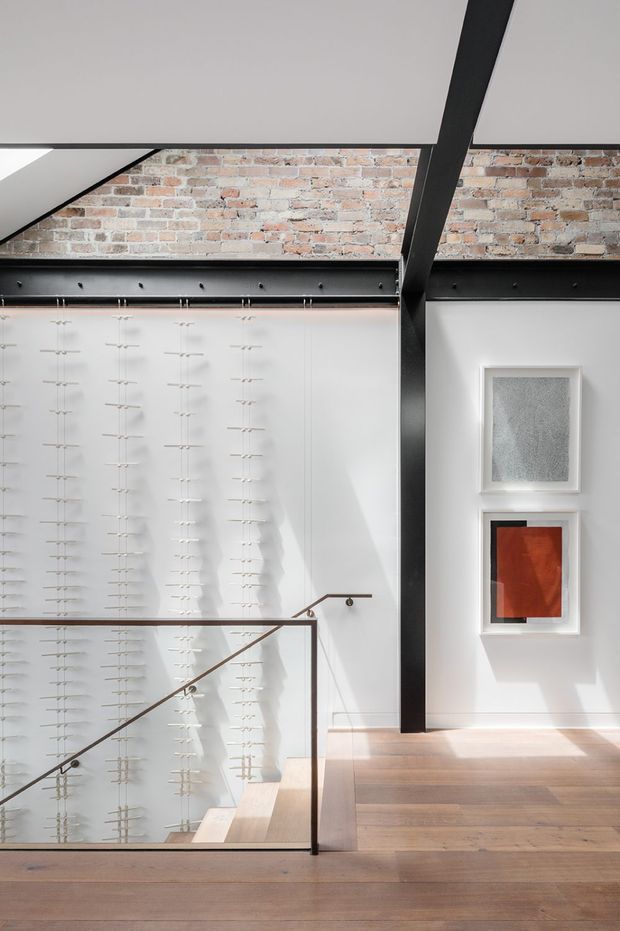 A bright, double-height industrial-style interior with exposed red brickwork, bold black steel beams, and a tall white wall featuring a large grid-like installation. A timber stair landing with a slim dark handrail and clear glass balustrade occupies the foreground, while two framed artworks hang on the right wall. Timber flooring and strong natural light create a refined gallery-like atmosphere.