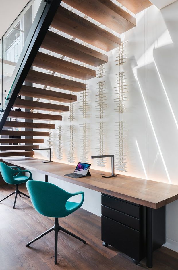 A bright, architecturally detailed home office with a long built-in timber desk beneath an open-riser staircase. The staircase features thick dark wood treads and a black structural stringer, while the white wall behind the desk has repeated vertical decorative timber elements. Two teal swivel chairs, slim black task lamps, laptops, and black drawer cabinetry furnish the workspace. Strong daylight enters through glazing and creates diagonal shadows across the white walls.
