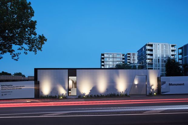 Twilight exterior view of a contemporary urban development, featuring a low rendered boundary or sales pavilion wall with illuminated landscaping and signage, backed by several multi-storey apartment buildings with extensive glazing and balconies. The image includes light trails from traffic along the street in the foreground and a clear blue evening sky.