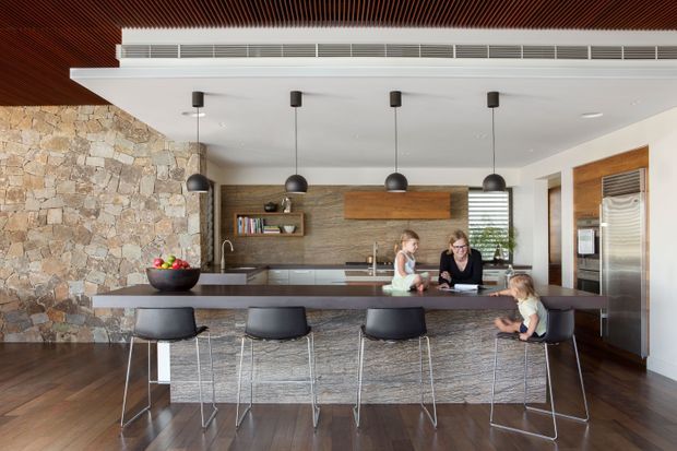 A spacious contemporary kitchen with a large island and four black bar stools. The room features a full-height irregular natural stone feature wall, dark timber flooring, a textured stone island front, dark wood ceiling slats, white cabinetry and walls, stainless-steel appliances, and four black pendant lights. A family is gathered around the island, with a dining or living area visible beyond the kitchen.