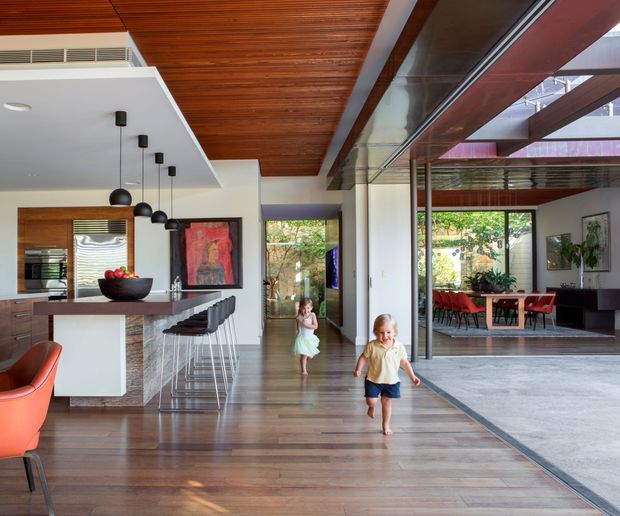 Bright open-plan contemporary home interior showing a kitchen island with stools on the left, a spacious living area and dining room visible through full-height glazing on the right, and a timber-lined ceiling with exposed beams. Warm timber flooring, white walls, dark-framed glazing, pendant lights, and large openings create a connected indoor-outdoor feel.