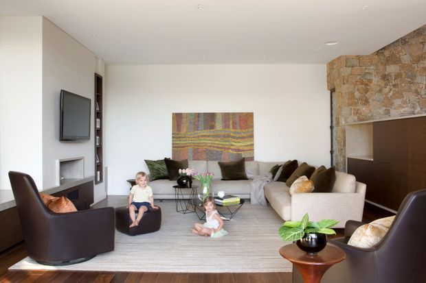 Bright contemporary living room with a large pale sectional sofa, dark lounge chairs, a wall-mounted television, built-in shelving, a linear fireplace, and a prominent rough stone feature wall. A colourful woven artwork hangs above the seating area, with a large neutral rug over dark timber flooring and a low coffee table. Two children and indoor plants are visible.