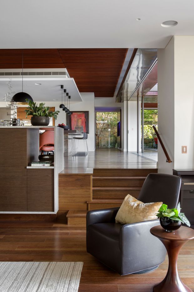 A refined open-plan interior viewed from a lower living area toward a raised kitchen and dining zone. The space features extensive dark timber flooring, a matching timber ceiling, a raised platform reached by timber steps, dark built-in kitchen cabinetry with a pale benchtop, pendant lighting, full-height glazing, and a black leather armchair beside a sculptural wooden side table. The composition is warm, contemporary, and strongly connected to the garden through the glazing.