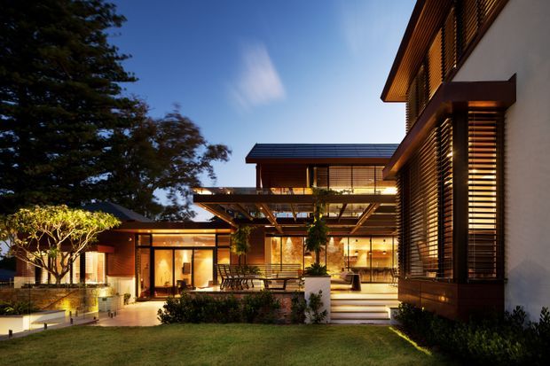 Twilight exterior view of a sophisticated multi-level contemporary residence. The composition features warm illuminated interiors, extensive timber horizontal louvres and screens, projecting roof planes, broad glazing, covered outdoor living areas, landscaped gardens, and a stepped terrace connecting the building wings.