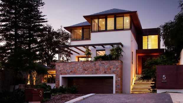 Exterior twilight view of a contemporary multi-storey residence with broad low-pitched hipped roof forms, illuminated horizontal louvre screens, white rendered walls, a natural stone-clad garage volume, timber-toned entry elements, and a landscaped driveway. A pergola projects over the upper-level outdoor area, while illuminated steps lead to the entrance.