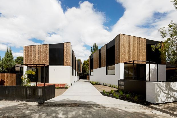 Street-facing view of a contemporary multi-unit residential development with a central concrete pedestrian driveway or walkway. The buildings feature crisp white rendered lower walls, dark charcoal upper volumes, extensive vertical timber cladding, black-framed glazing, and low landscaped garden edges. Strong geometric massing and contrasting natural materials create a modern architectural composition.