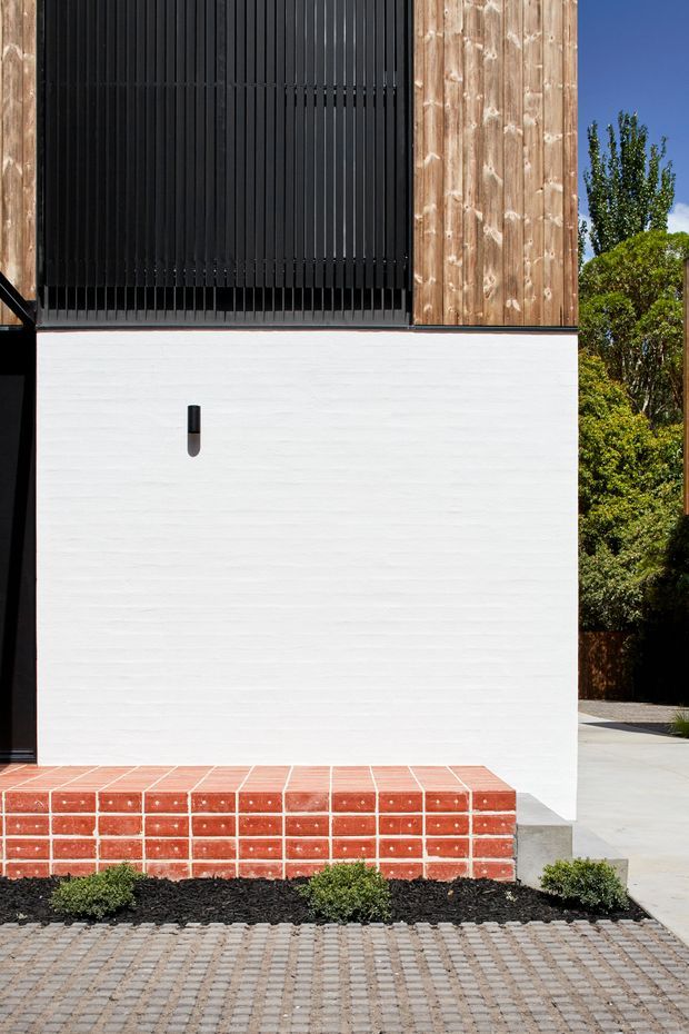 Close-up exterior view of a contemporary residential facade. The composition features a crisp white rendered masonry wall, a projecting red brick-tiled step or plinth, vertical timber cladding, and a large black vertically screened opening or window. A neat landscaped strip with low shrubs and dark mulch borders a paved driveway or walkway in the foreground.