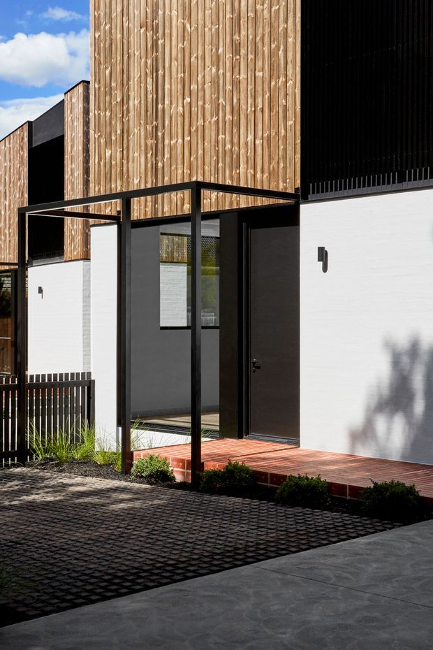 A contemporary residential facade viewed from the driveway, featuring strong geometric forms, vertical timber cladding, white rendered walls, dark metal-framed entry glazing, and a flat-roofed black steel canopy over a raised red-tiled entrance terrace. The foreground includes dark patterned paving and a simple landscaped edge.