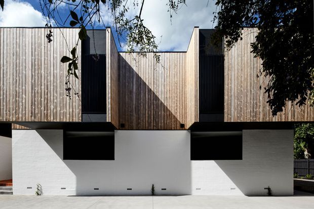Front exterior view of a contemporary multi-level residential building with a strongly geometric composition. The upper facade is clad in vertical natural timber boards, interrupted by dark recessed window openings and projecting box-like volumes. The lower level has smooth white rendered walls with deep black shadowed recesses and sharp angular sunlight patterns. A paved forecourt and partial surrounding foliage are visible.
