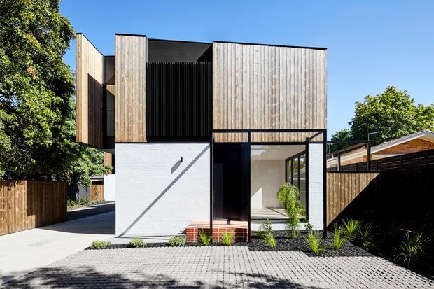 Exterior view of a contemporary multi-level residence with a strongly geometric composition. The facade combines vertical light timber cladding, dark recessed panels, white rendered masonry, and extensive black-framed glazing. A paved forecourt, timber boundary fencing, planted garden beds, and a partially visible pitched-roof structure frame the house.