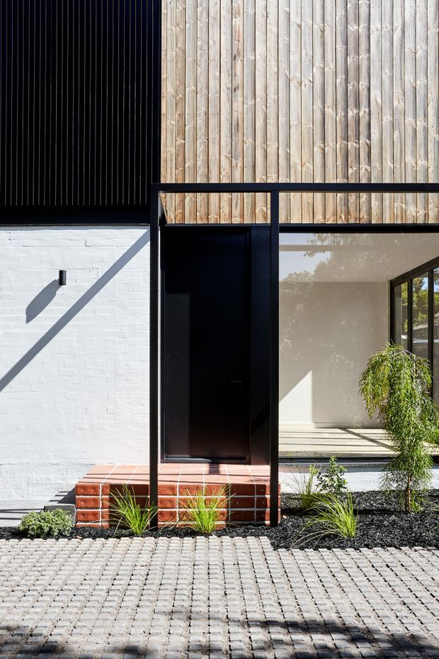 A contemporary residential facade viewed from the street, combining a black vertically ribbed upper wall, pale vertical timber cladding, and a white-painted brick lower wall. A tall black entry door sits beside large glazing, with a red-brown tiled entry step, dark landscaped garden bed, grasses, and a light grey cobblestone-paved foreground.