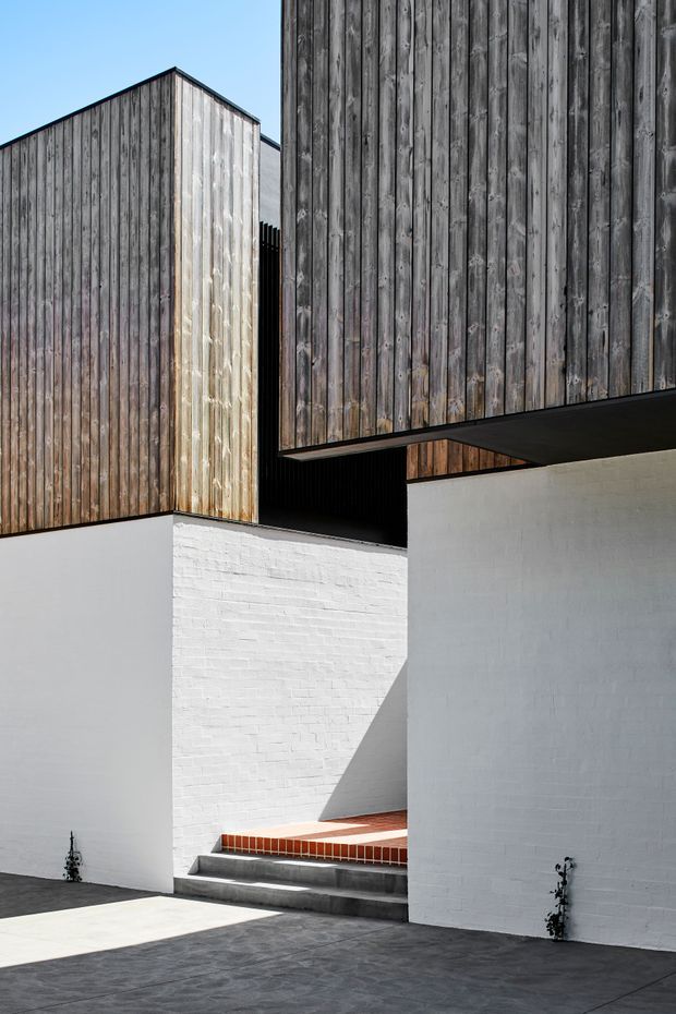 Minimalist contemporary exterior architecture featuring intersecting cubic volumes, weathered vertical timber cladding, white-painted brick and plaster walls, a projecting dark soffit, and a stepped entry courtyard with concrete steps and terracotta brick paving. Strong sunlight creates geometric shadows across the restrained material palette.