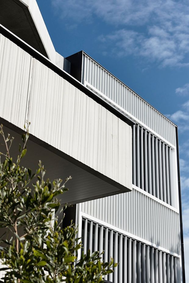 Upward-looking close view of a contemporary multi-storey building facade with white vertically ribbed cladding and prominent vertical louvre screens. Strong geometric projections and recessed openings create layered shadow lines against a blue sky, with some foreground greenery at the base.