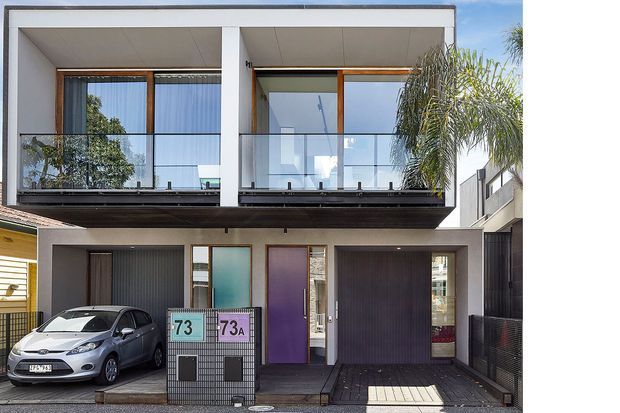 Street-facing view of a contemporary multi-storey urban residence with a strong rectangular facade, upper-level balconies enclosed by clear glass balustrades, timber-framed glazing, white rendered walls, dark tiled lower facade elements, and two ground-floor garage doors. A compact driveway and parked car are visible in front, with neighbouring buildings closely surrounding the property.