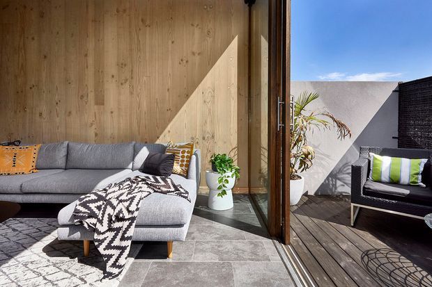 A bright contemporary indoor-outdoor living area with a large grey sectional sofa, patterned cushions and throw, natural timber wall panelling, grey stone-look flooring, and full-height glazed timber doors opening onto a timber deck and small outdoor seating area. Strong sunlight creates geometric shadows across the warm wood and neutral finishes, with potted plants adding greenery.