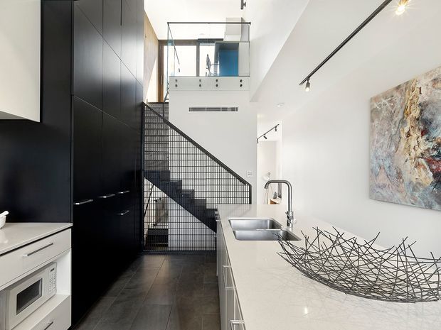 A contemporary open-plan kitchen viewed toward a black metal mesh staircase. The kitchen features a long pale stone island with integrated double sink and gooseneck tap, white lower cabinetry, tall matte-black cabinetry, dark tiled flooring, and track lighting. A mezzanine or upper-level void is visible above the stair, with an abstract artwork on the adjacent white wall and a decorative wire bowl on the island.