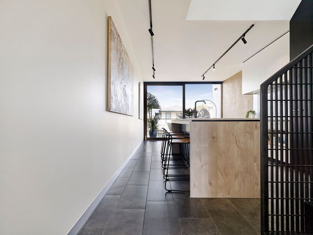 Bright contemporary kitchen with a compact timber island and integrated seating, dark large-format tiled flooring, white walls, black track lighting, and a black vertical-slat stair or screen partition. A large rear window brings in daylight and overlooks neighbouring buildings. The space has a clean, restrained palette with light timber and dark accents.