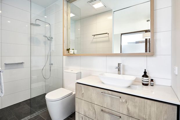 Bright contemporary bathroom with a frameless glass shower enclosure, wall-mounted shower fittings, white tiled walls, dark grey tiled floor, compact toilet, and a floating light timber vanity with a white vessel basin. A large timber-framed mirrored cabinet spans the vanity wall, with a small horizontal window above the basin.