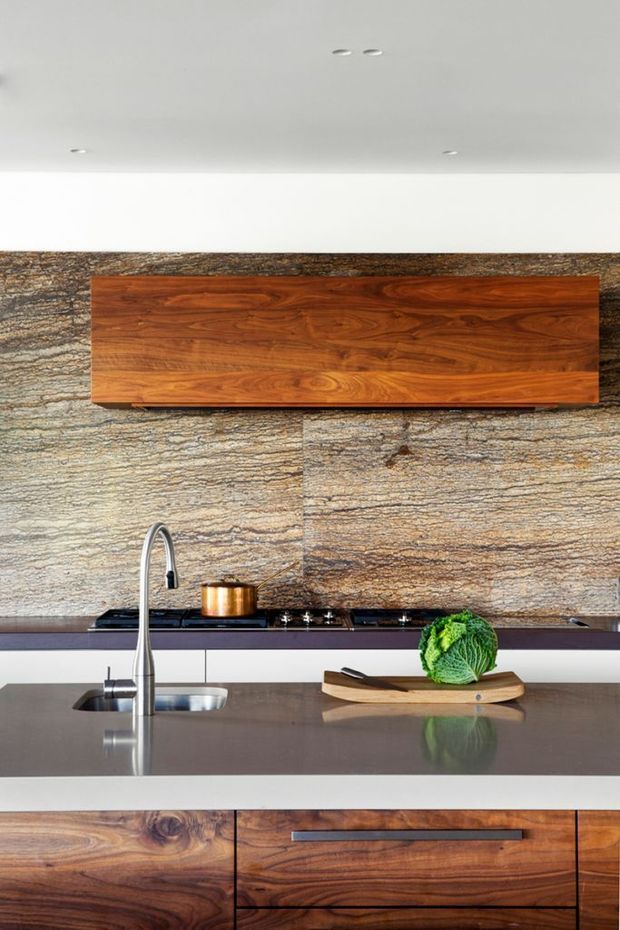 Close-up view of a contemporary kitchen with a large grey stone island benchtop, integrated stainless-steel sink and curved tap, gas cooktop, and dark cabinetry. The backsplash is clad in horizontally textured natural stone, complemented by warm walnut timber cabinetry and a matching wall-mounted feature cabinet. Recessed ceiling lights and minimalist detailing create a refined, modern fit-out.
