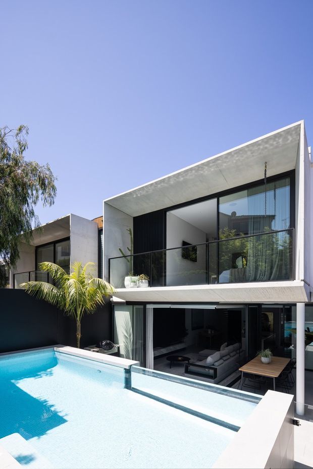 A contemporary multi-level residence with crisp white geometric concrete forms, extensive black-framed glazing, and glass balcony balustrades overlooking a bright swimming pool. The open lower level reveals a living area and outdoor dining zone, while a palm and surrounding planting soften the angular architecture.