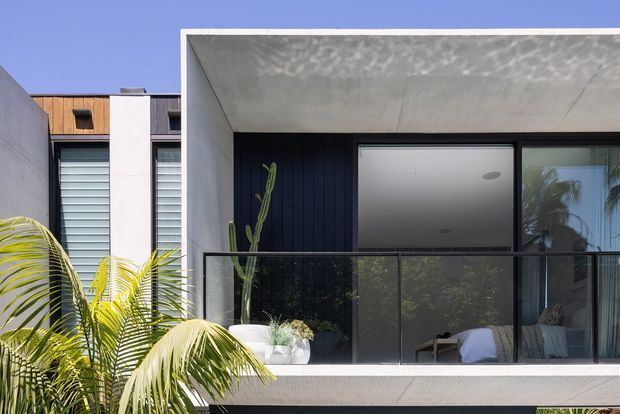 Exterior view of a contemporary multi-level residence with a projecting rectangular concrete-framed upper balcony or terrace. The facade combines smooth white rendered/concrete surfaces, dark vertical cladding, large glazed openings, and black-framed glass balustrading. Tropical palms and potted plants soften the modern composition, while bedroom furnishings are visible through the glazing.