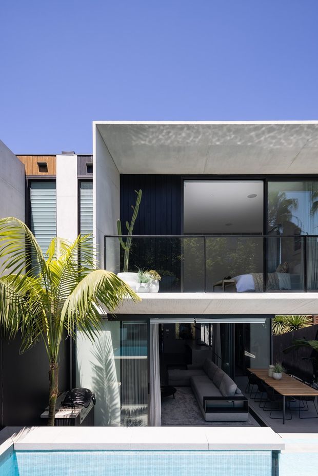 Contemporary two-storey residential facade with a projecting concrete balcony and deep overhang, dark vertical cladding, large glazed openings, and a black-framed glass balustrade. A tropical palm borders the outdoor entertaining area, with a swimming pool in the foreground. Through the lower glazing, a living room and outdoor dining area are visible.