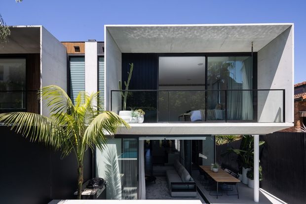 Contemporary two-storey residential facade with a strong box-like composition, deep upper-level concrete-framed balcony, extensive dark-framed glazing, and a lower outdoor living area. A palm tree and potted plants soften the monochrome architecture, while interior living furniture is visible through the ground-floor glazing.