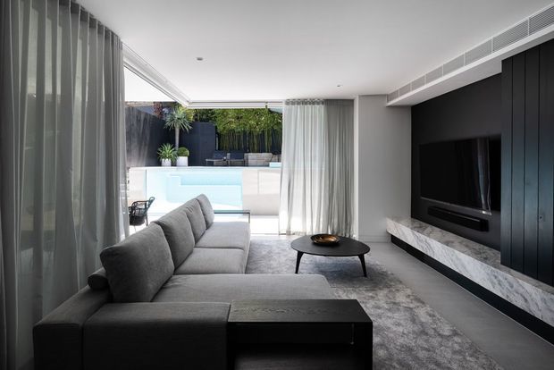 A refined contemporary living room with a large grey sectional sofa, black oval coffee table, and wall-mounted television set into dramatic black vertical-panel joinery. A long marble-look hearth or media console runs beneath the television. Full-height sheer curtains soften the glazing on the left and frame an open connection to a bright outdoor pool courtyard, with integrated linear air-conditioning grilles along the ceiling. The palette is monochromatic grey, white, and black, creating a calm, sophisticated atmosphere.