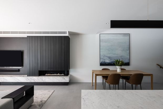 Contemporary open-plan living and dining area with a minimalist built-in fireplace and media wall, vertical dark timber panelling, pale stone hearth, light flooring, a timber dining table with upholstered chairs, and a large pale stone kitchen island in the foreground. A muted ocean-toned artwork and simple plant provide restrained decorative accents.